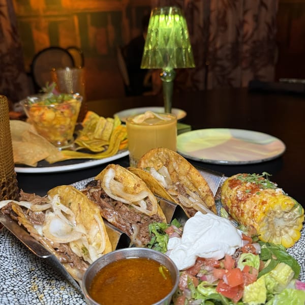 A plate of tacos, elote, salad, and dipping sauce, with chips, salsa, and a drink in the background.