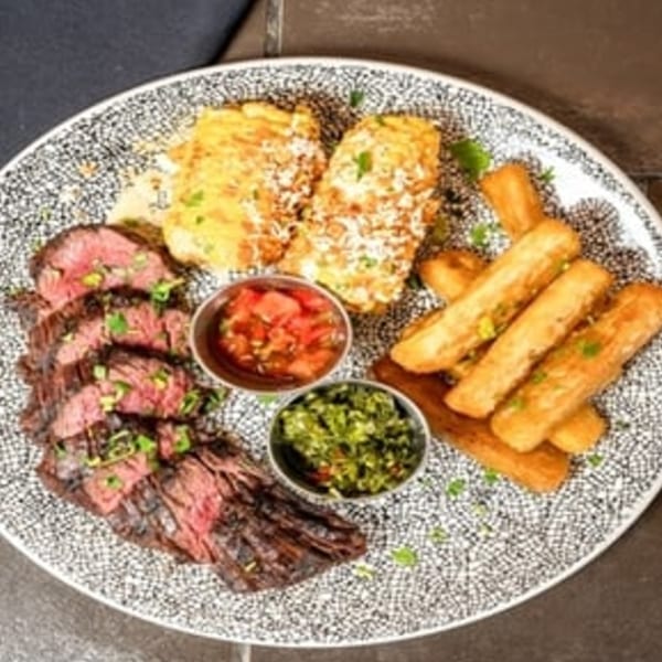 Sliced steak, two corn on the cob, yuca fries, and two dipping sauces on a patterned plate.