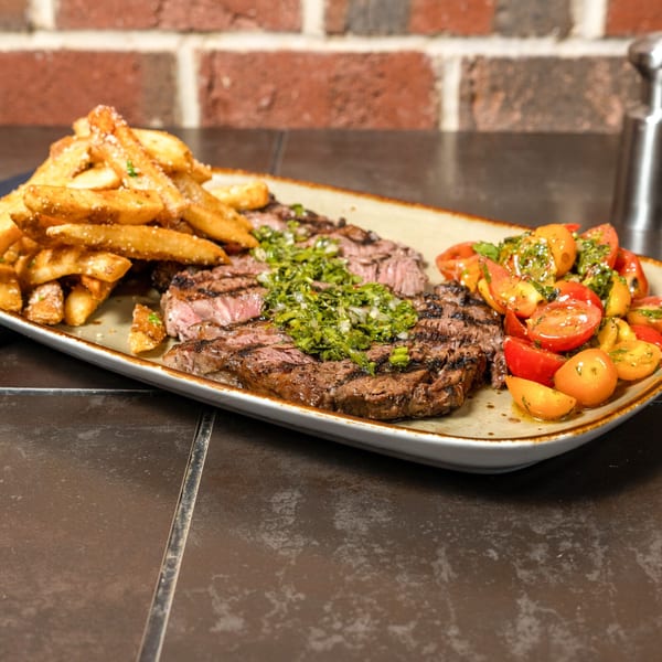 Grilled steak with chimichurri, a side of fries, and a tomato salad on a rectangular plate.