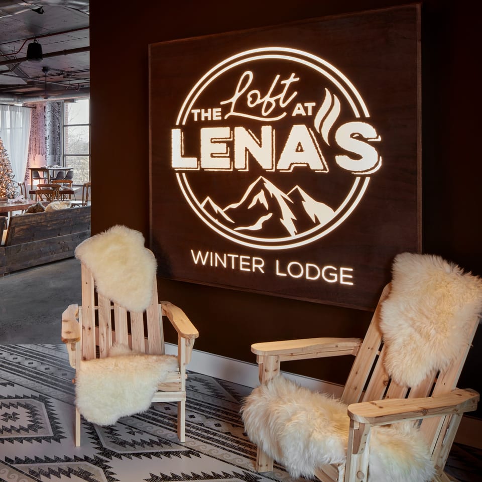 Two wooden Adirondack chairs with fluffy white throws sit before a glowing "The Loft at Lena's Winter Lodge" sign.