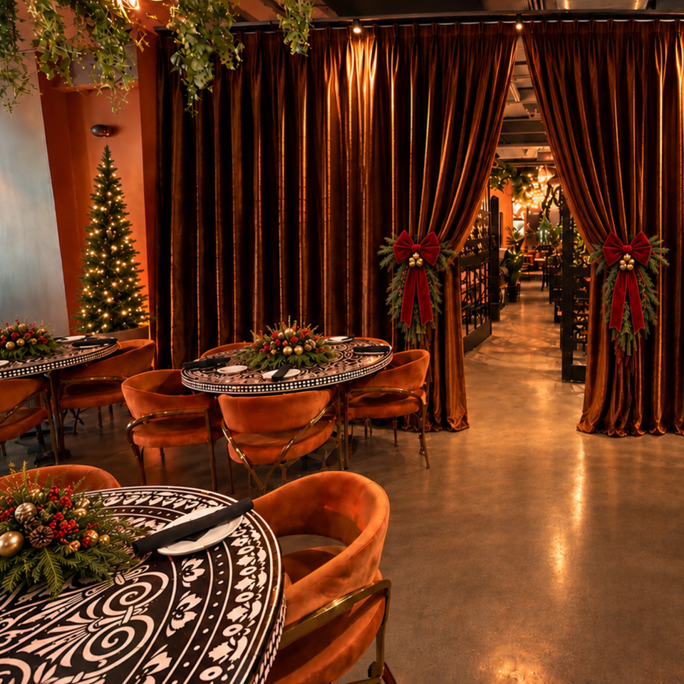 Warmly lit restaurant dining room with patterned tables, orange chairs, velvet curtains, greenery, and festive Christmas decorations.