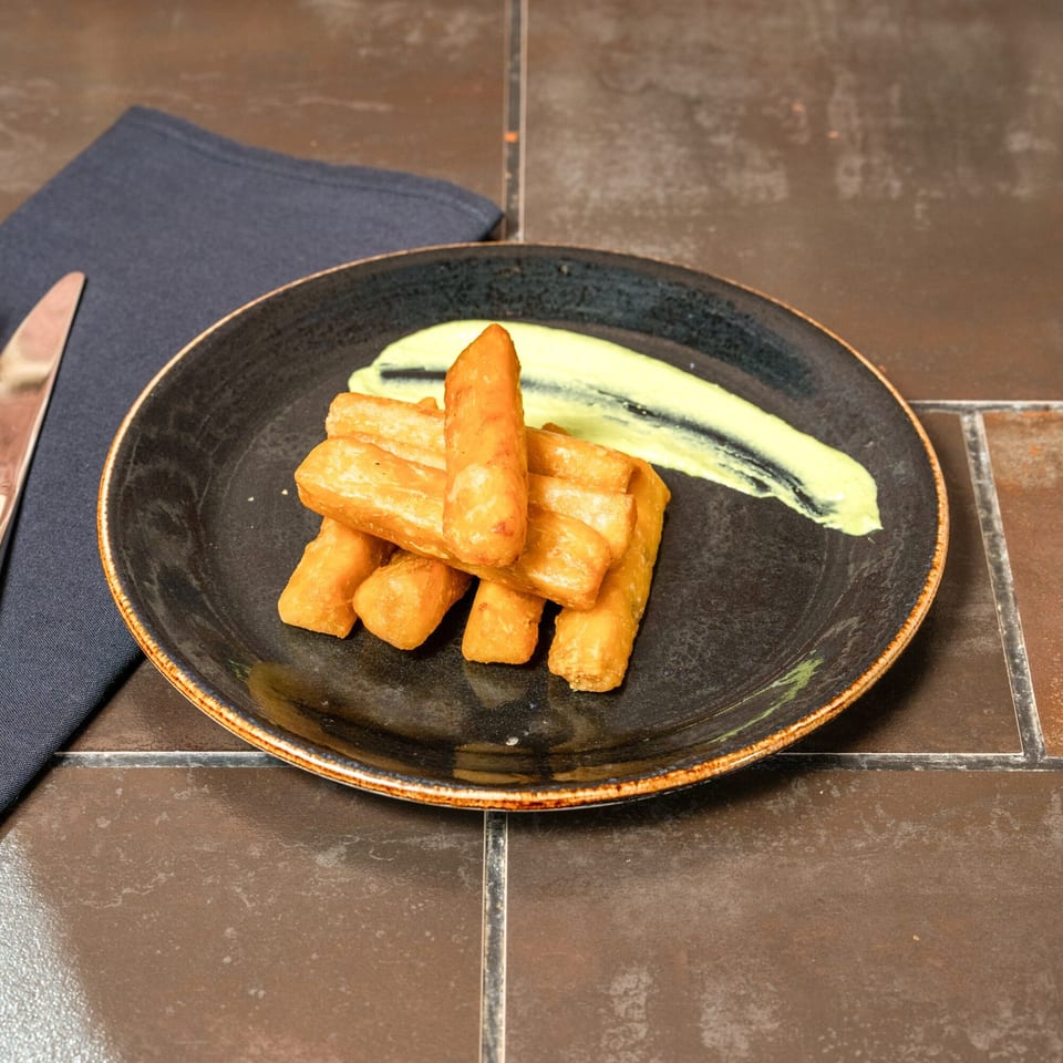 Golden-brown yuca fries with a swirl of light green sauce on a dark plate, next to a napkin and knife.