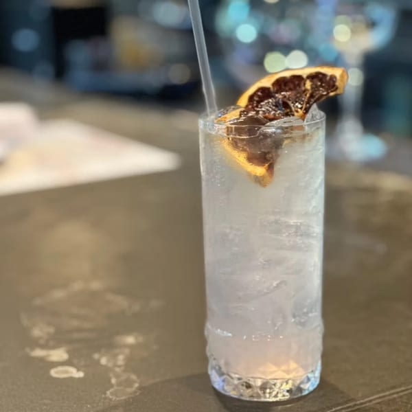 A tall glass of clear, iced cocktail with a dried orange slice and straw on a bar.