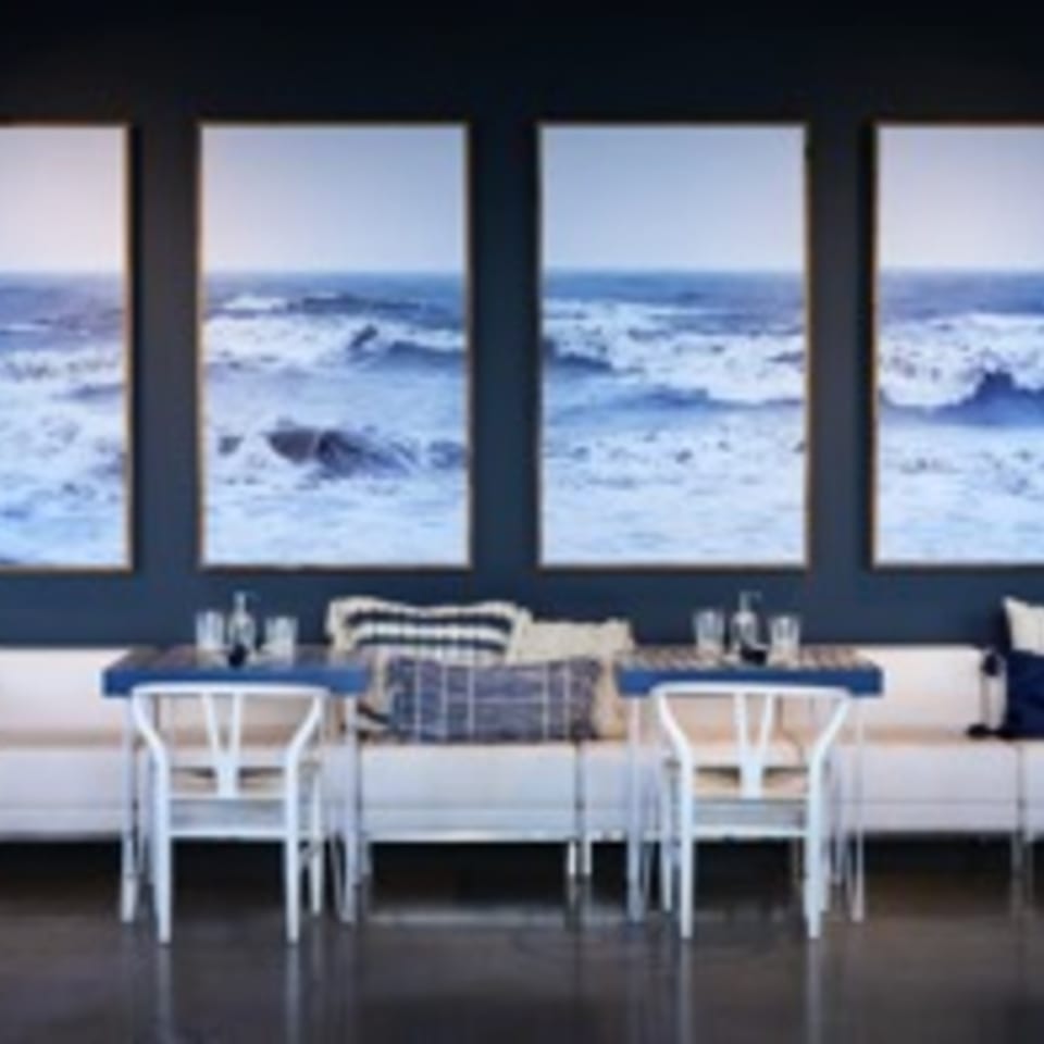 Dining area with white banquette seating, blue tables, and large ocean wave art on a dark blue wall.