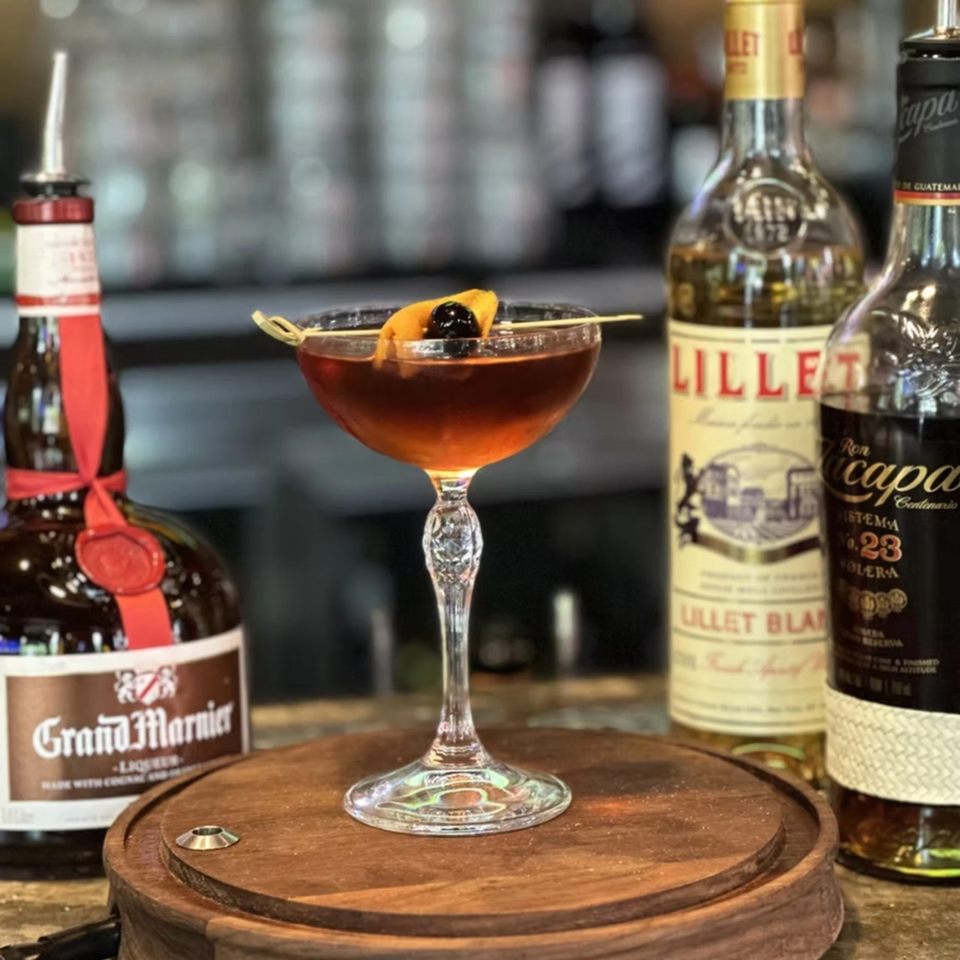 A Cuban cocktail garnished with orange peel and cherry, surrounded by liquor bottles on a wooden tray.