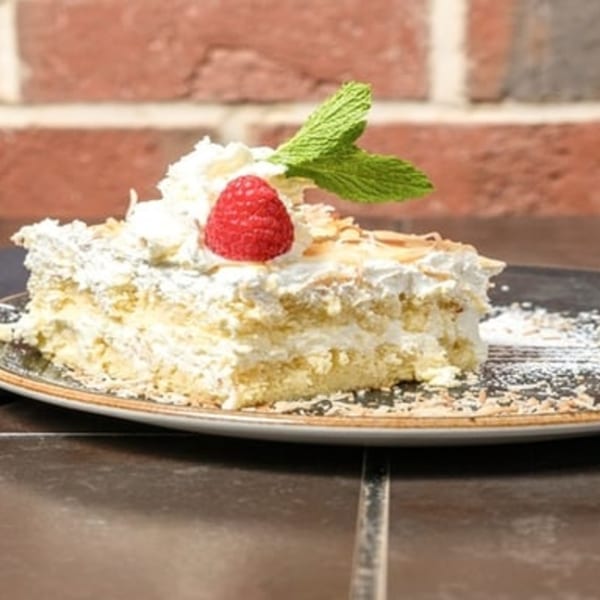 Slice of layered cake with white frosting, topped with a raspberry and mint, on a dark plate.